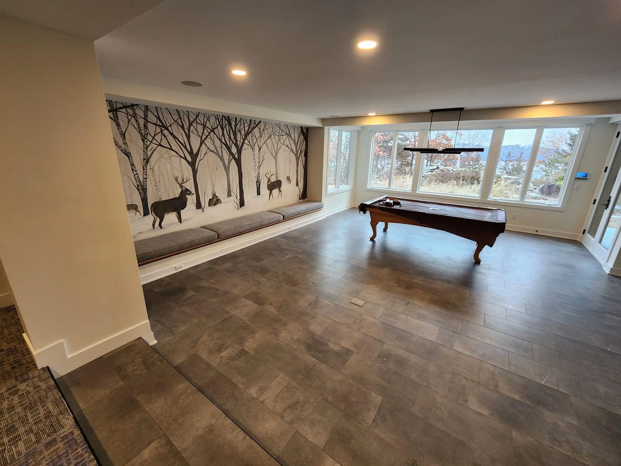 Modern recreation room with LVP flooring, built-in window seat, forest mural, and pool table