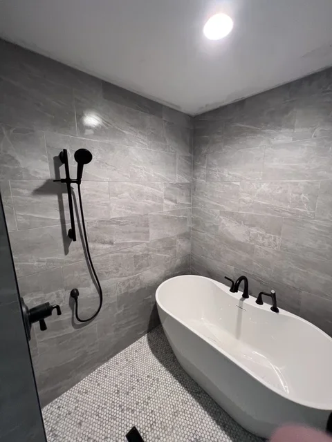 Modern bathroom with gray tile walls, white soaking tub, and black fixtures