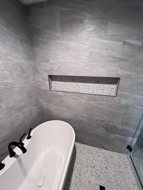 Modern bathroom with gray tile walls, white soaking tub, and frameless glass shower enclosure